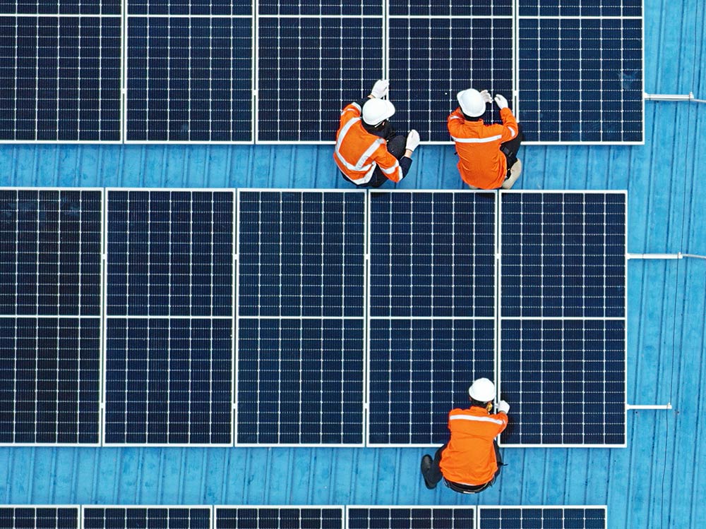 Three men in orange hi-visibilty jackets installing solar panels on a blue tin roof.